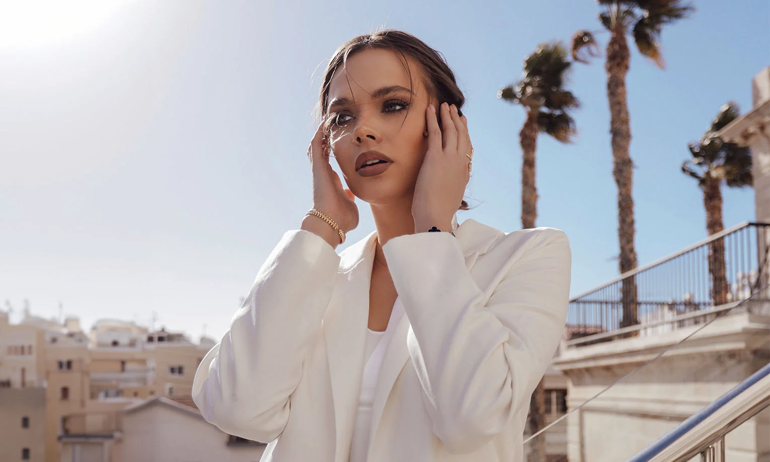 A stylish woman outdoors wearing a white blazer, with her hands touching her face, and a sunny background with palm trees - Botox and Wrinkle Relaxers in West Hollywood, CA