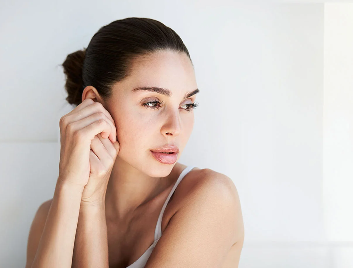 A brunette woman with glowing skin and neatly tied-back hair, resting her hand gently near her ear in a bright, minimalistic setting - Facial Fat Grafting in West Hollywood, CA