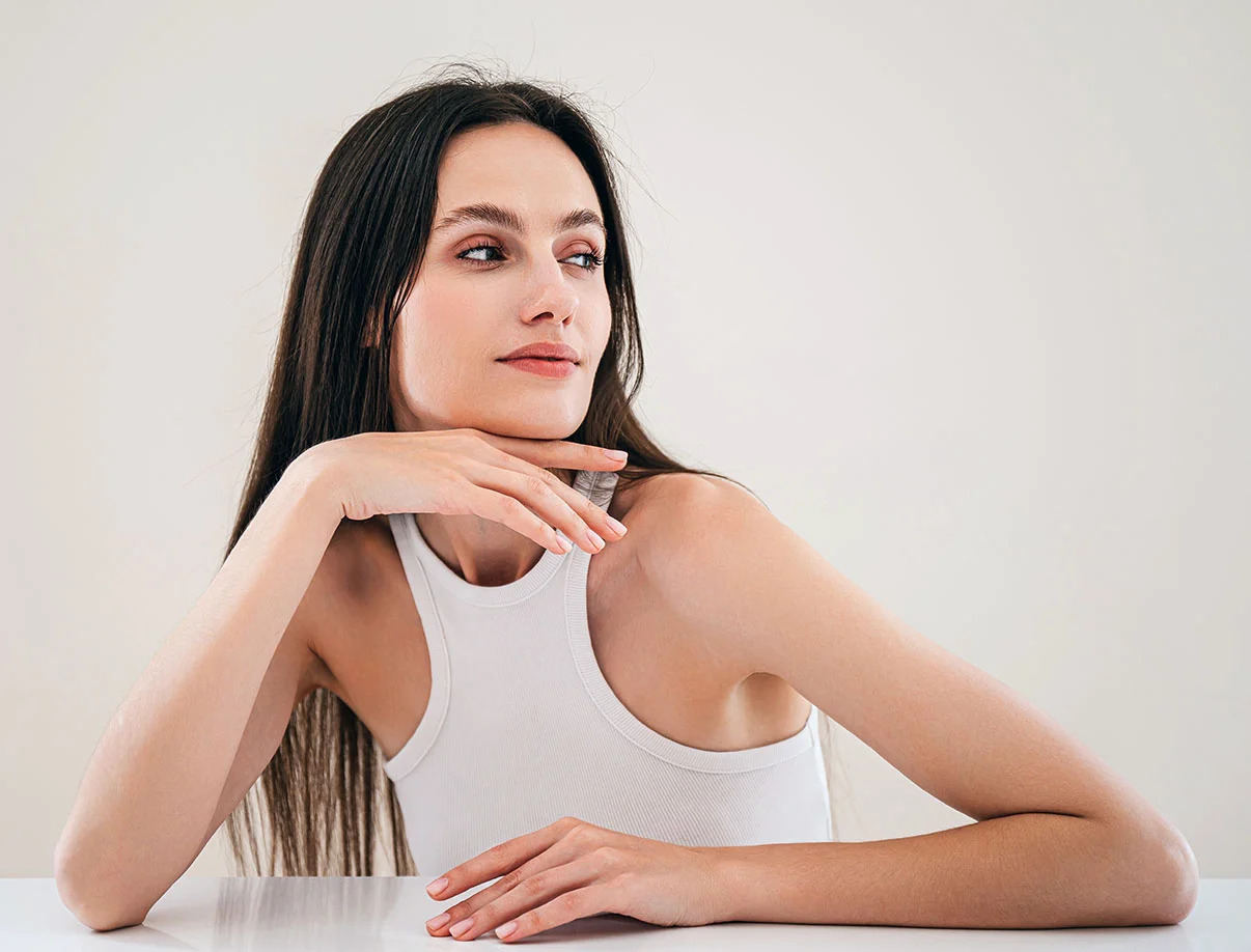 Elegant woman in a white tank top, resting her chin on her hand and looking away thoughtfully - Facial Reshaping in West Hollywood, CA