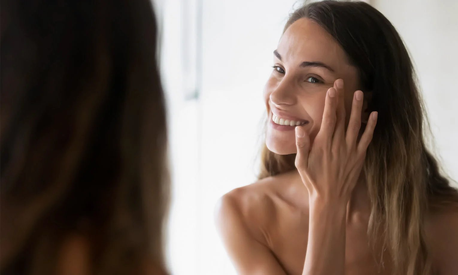 Close-up of a happy woman gently touching her cheek while looking in the mirror, showcasing natural beauty - hydrafacial in West Hollywood, CA