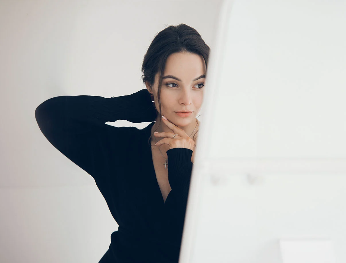 Young woman dressed in black with a simple necklace, thoughtfully adjusting her hair in front of a lighted mirror - microneedling in West Hollywood, CA