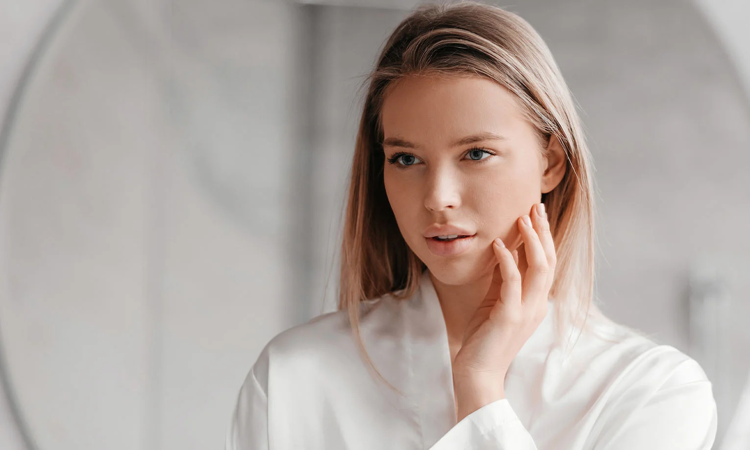 A young woman in a white robe gently touches her face, gazing thoughtfully, with a soft-focus background of muted colors. - PRFM PRP EZ Gel in West Hollywood, CA