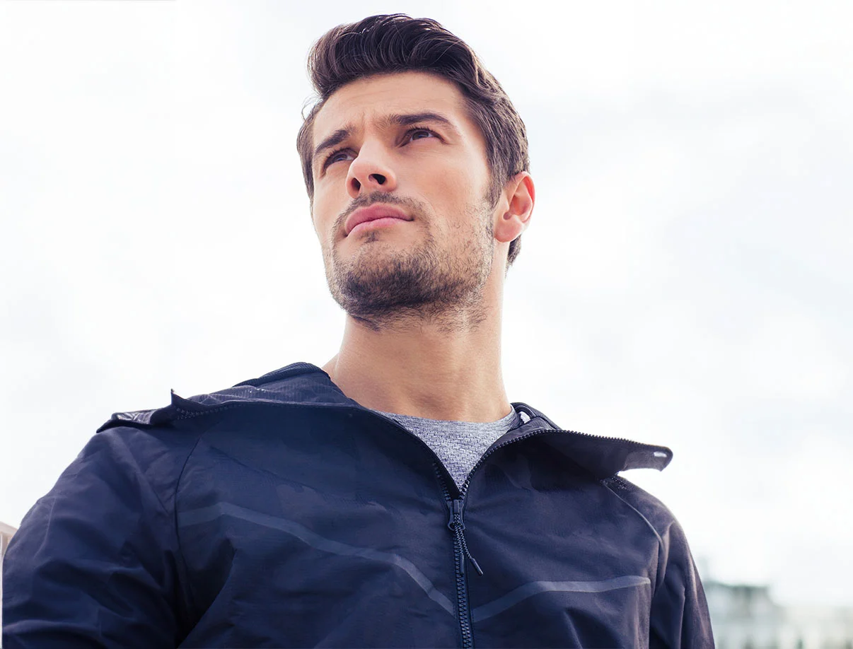 Athletic-looking man outdoors wearing a black jacket, looking off into the distance with a focused expression under a bright sky - PRP Hair Restoration in West Hollywood, CA