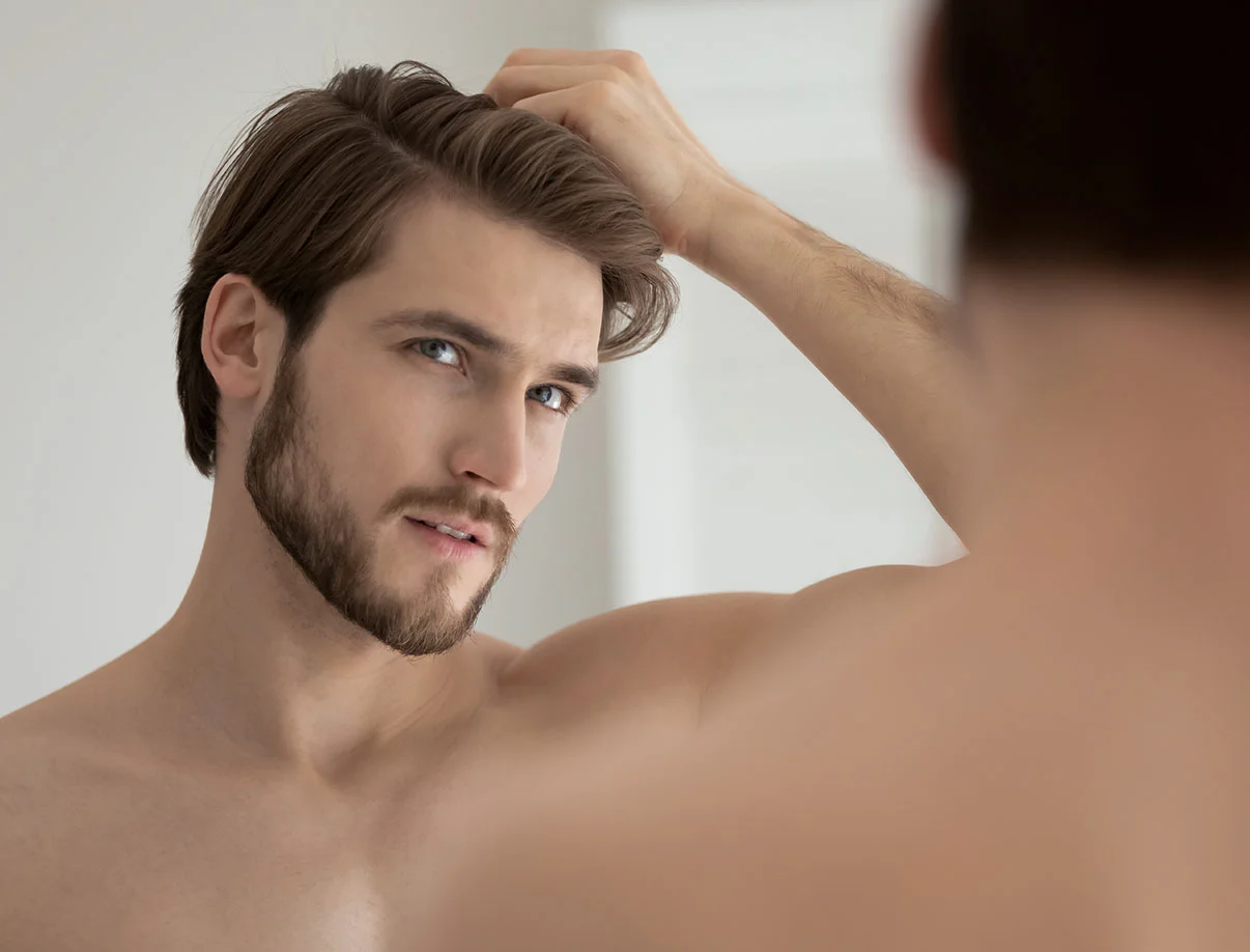 Young man examining his hairline in a mirror, with a contemplative expression in natural lighting - Smart Graft Hair Restoration in West Hollywood, CA