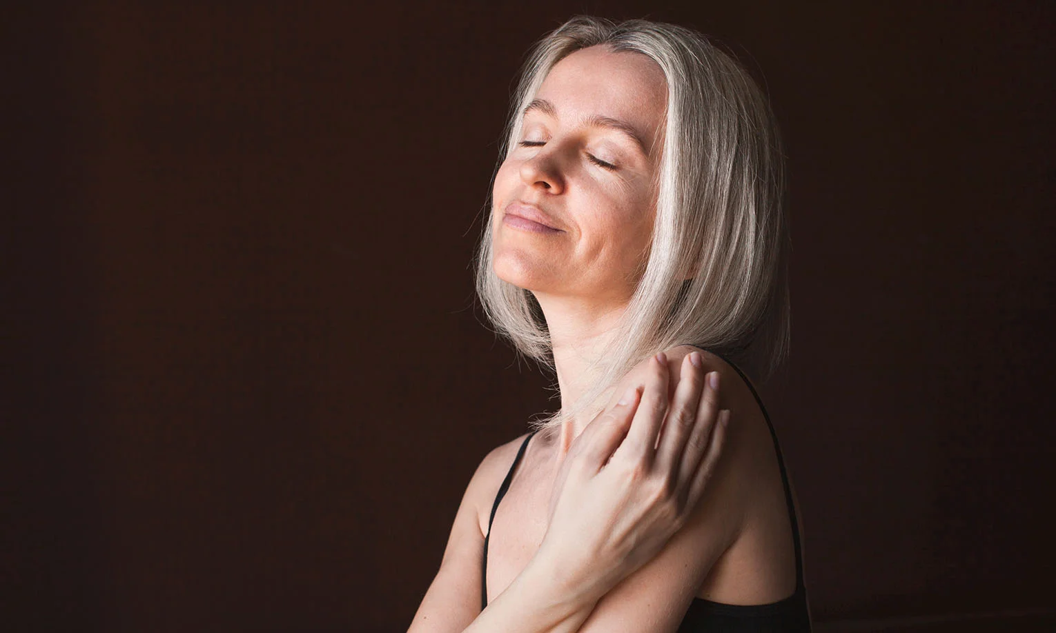 Close-up of a woman with silver hair and a serene expression, gently holding her shoulder against a dark backdrop - Vitamin Shots in West Hollywood, CA